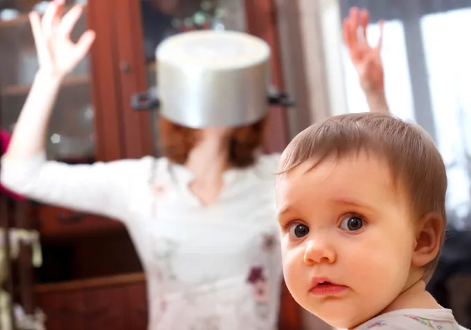 Mother has a pot over her head and seems frustrated with baby. Baby looks confused - good parenting