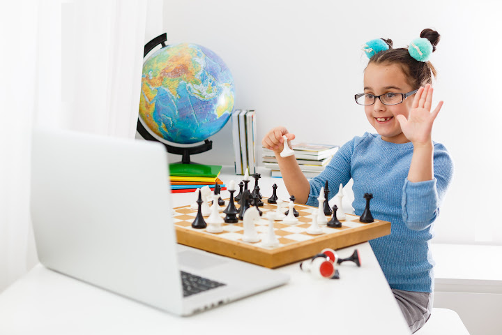 girl raises hand playing chess in front of a laptop