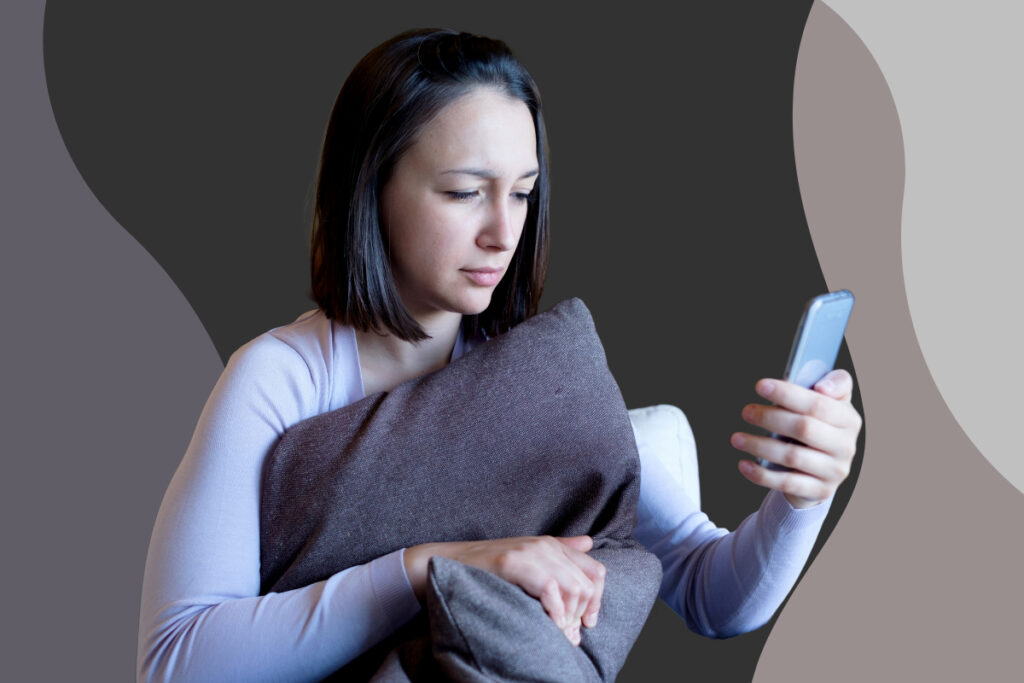 A woman hugs a pillow and stares at her phone.