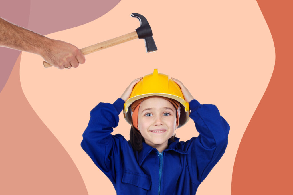A young girl wearing a safety hat. An adult's hand above her head is holding a hammer.