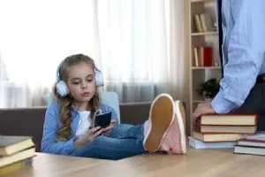 girl with headphones and feet on desk, dad stands over giving her natural consequences